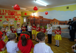 Dzieci z gr. IV: dziewczynki w czerwonych podkoszulkach i żółtych spódniczkach oraz chłopcy w białych koszulach i czarnych spodniach ze skrzyżowanymi rękoma recytują wiersz.